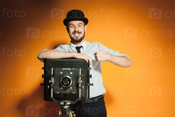 Young  positive  smiling man in hat as photographer with retro camera on an orange background