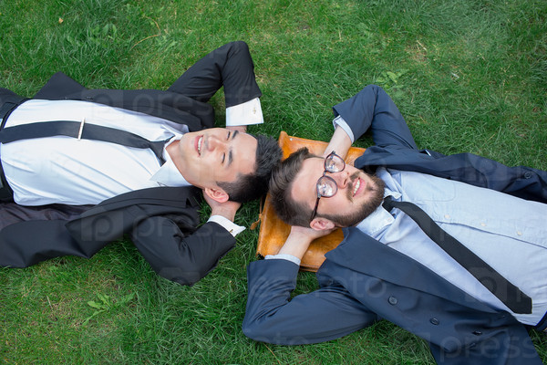 The two happy young businessmen in a suites lying on the green grass, top view. The one man is European, other is Chinese