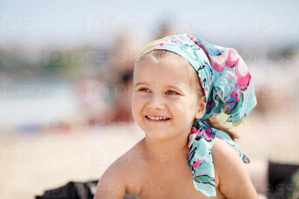 A smiling little girl child with a colorful handkerchief on his head. Hot sunny summer day at the beach. Selective focus on child\'s face.