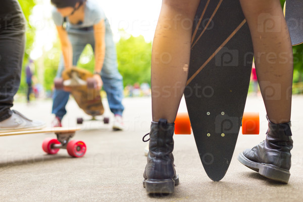 Teenagers practicing long board riding outdoors in skateboarding park. Detail of legs of girl wearing black boots and stockings holding long board in foreground. Active urban life. Urban subculture, stock photo
