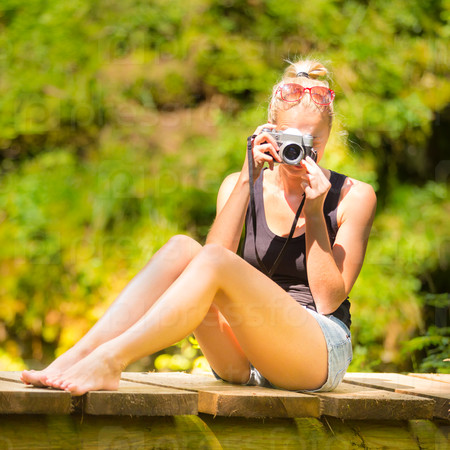 Beautiful barefooted blonde caucasian girl wearing jeans shorts an sporty black sleeveless t-shirt, sitting on a vintage wooden bridge, taking photos with retro camera.