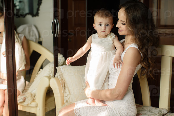 Portrait of happy young attractive mother playing with her baby girl near window in interior at haome. Pink dresses on mother and