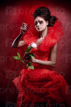 Red Queen. Woman with creative make-up in fluffy red dress with a white rose and paintbrush posing indoors