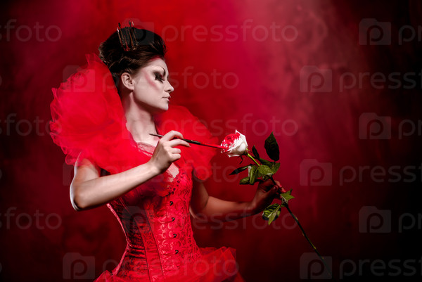 Red Queen. Woman with creative make-up in fluffy red dress with a white rose and paintbrush posing indoors