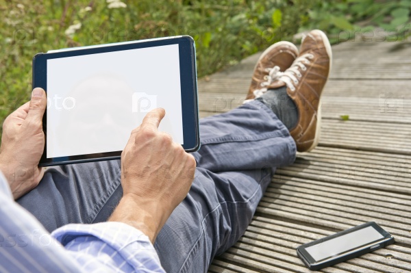 Man, tapping on a tablet, playing a game outdoors, sitting on a plank bridge, whith his phone next to him. Both can be used as presentation for mock-up apps, website and presentations.