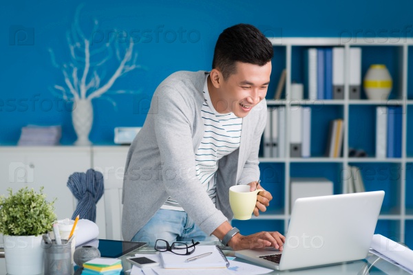 Young man with a cup of coffee laughing while reading something on laptop