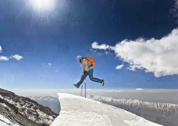 happy man with orange backpack and two poles jumpingon a cliff in mountains at sunrise