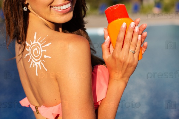 Sexy smiling girl with sun shape from sunscreen lotion on her shoulder is resting near swimming pool with blue water, summer time