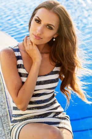 Sexy smiling woman is wearing striped fashion dress with tanned skin and makeup is sitting near pool