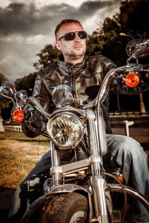 Biker man wearing a leather jacket and sunglasses sitting on his motorcycle looking at the sunset.