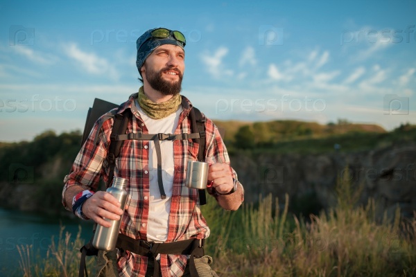 Young caucasian tourist with backpack resting and drinking water on the top of hill, stock photo