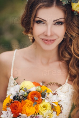 Bride in blooming apple trees. Woman in flower wreath and wedding bouquet in hands. Beautiful bride in wedding dress posing in a blooming apple garden. Spring mood. Young woman outdoors