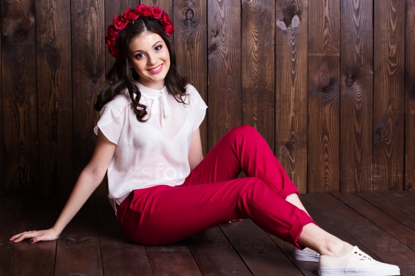 Fashion portrait of cute teenager girl with nice makeup is wearing white blouse and red roses wreath on her head sitting on wooden background