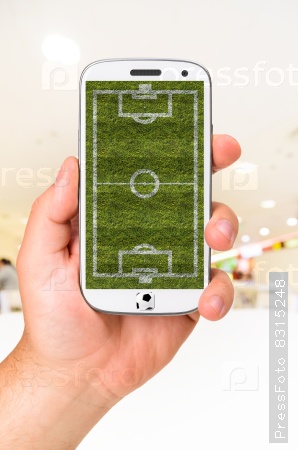 male hand is holding modern phone with soccer or football field on screen against blurred interior