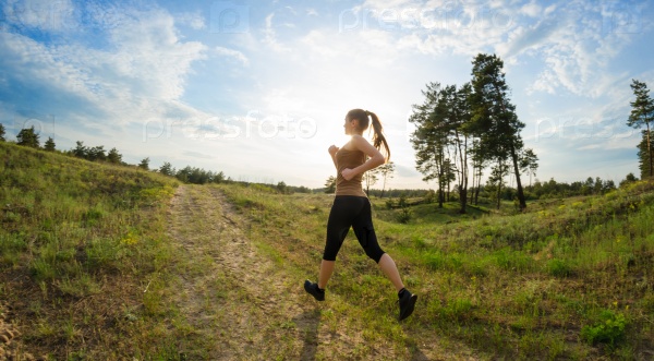 Young woman jogging outdoors. View from the back, on a background of blue sky with patches of sunlight and green field.