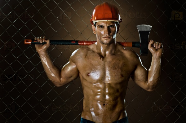 the muscular tired worker chopper man, in safety helmet with big heavy ax in hands, on netting fence background