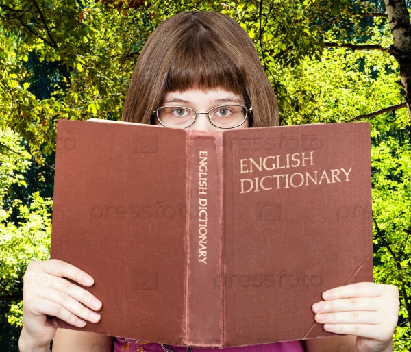 girl with spectacles looks over English Dictionary book with green summer forest on background