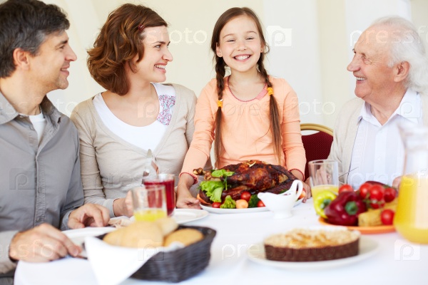 Happy family sitting at Thanksgiving table and looking at cute girl with roasted turkey