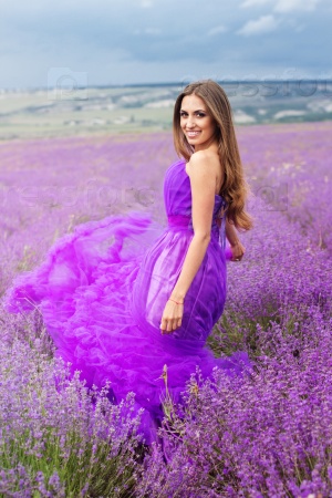 Beautiful smiling woman is wearing magic purple fashion dress posing at field of purple lavender flowers