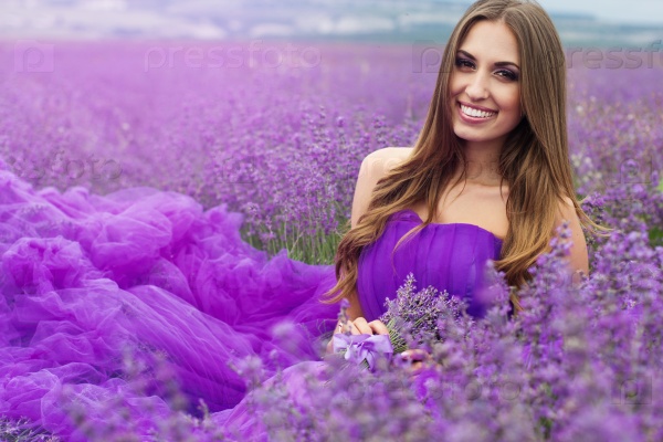 Beautiful smiling woman is wearing magic purple fashion dress posing at field of purple lavender flowers