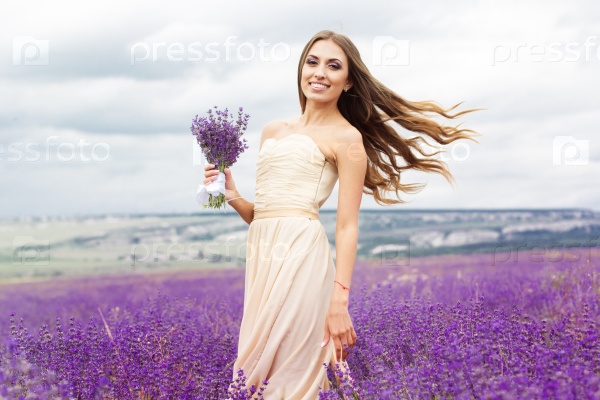 Beautiful smiling woman is wearing nice wedding dress playing with bouquet at field of purple lavender flowers