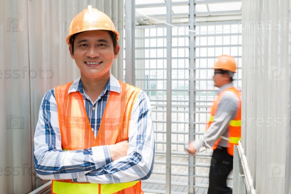 Portrait of Asian engineer in orange vest and hardhat smiling and looking at the camera