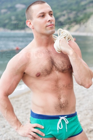 Sexy young guy body builder with six pack is posing on the beach with rope