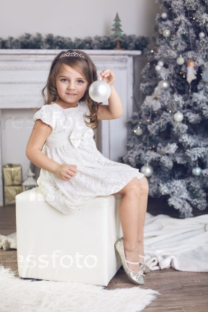 5 years old little girl dressed in beautiful fashion white flower dress holding toy and sitting near Christmas tree
