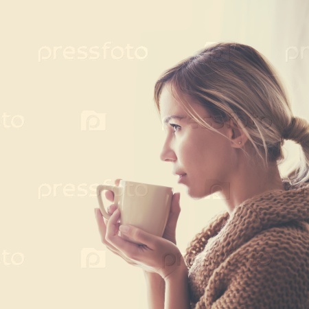 Woman wearing warm knitted sweater is drinking cup of hot tea or coffee near window in autumn morning sunlight, photo warm toned