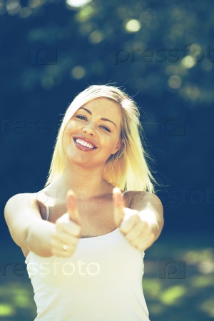 Young happy woman giving thumbs up sign.Blonde attractive girl with white sleeveless blouse on blurred  background  smiling to a camera.Shallow depth of field focus on face.