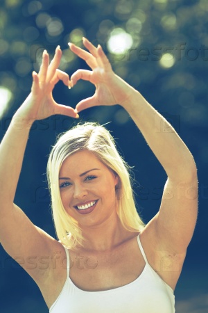 Young happy woman creates heart by her fingers.Blonde attractive girl white sleeveless blouse on blurred background in a park.Beautiful  Caucasian female model.Shallow depth of field focus on face.