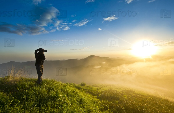 man with camera making photo on a hill at sunset and clouded sky