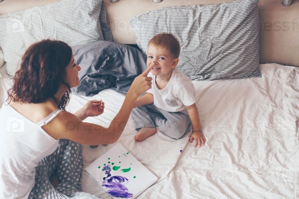 Young mother with her 2 years old little son dressed in pajamas are relaxing and playing in the bed at the weekend together, lazy morning, warm and cozy scene. Pastel colors, selective focus, stock photo