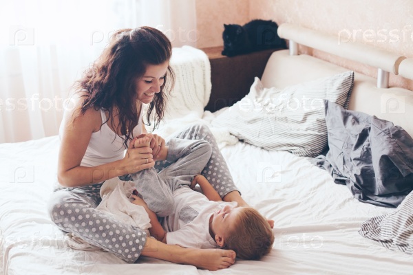 Young mother with her 2 years old little son dressed in pajamas are relaxing and playing in the bed at the weekend together, lazy morning, warm and cozy scene. Pastel colors, selective focus, stock photo