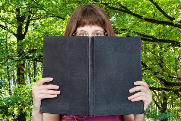 girl with glasses looks over big book with blank cover and green woods on background