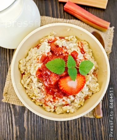 Oatmeal in a bamboo bowl with strawberry and rhubarb sauce on a napkin of burlap, spoon, milk in glass jug, rhubarb against the dark boards on top