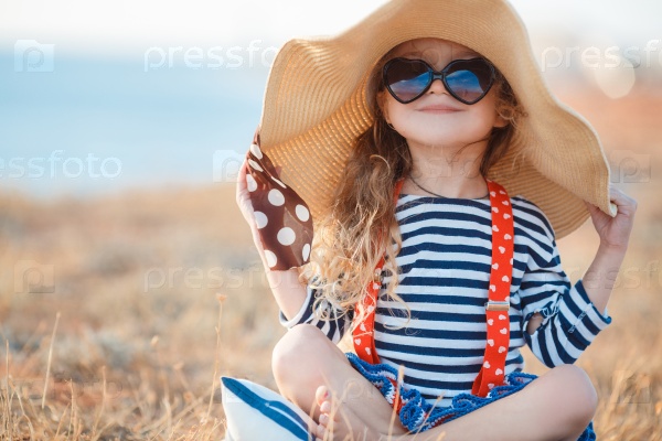 Pretty little girl in a striped dress and hat relaxing on the beach near sea, summer, vacation, travel concept. smiling cute little girl on beach vacation. Baby girl in hat and sun glasses on beach