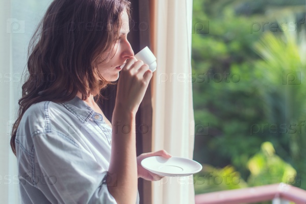 Woman drinking tea or coffe and looking through the window.  Young lady meeting sunrise