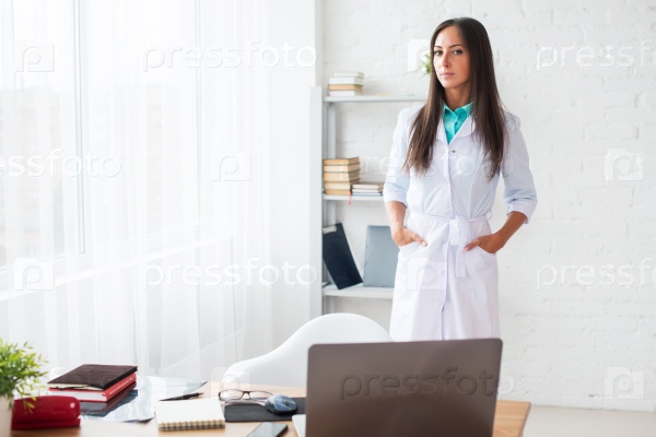 Portrait of young woman doctor with white coat standing in medical office looking at the camera.