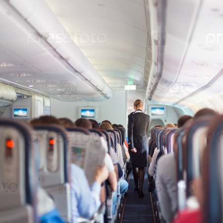 Interior of commercial airplane with passengers on seats during flight. Stewardess in dark blue uniform walking the aisle. Square composition, stock photo