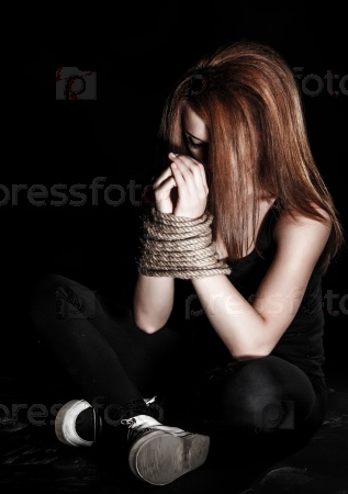 Beautiful young woman with tied arms sitting on the floor over black background