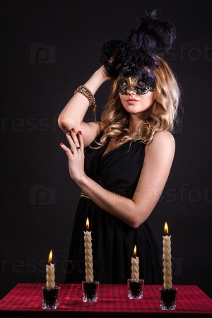 Mysterious woman in a mask near the table with alight candles over black background