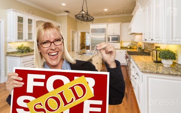 Happy Young Woman Holding Sold For Sale Real Estate Sign and Keys Inside Beautiful Custom Kitchen.