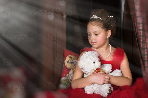 Thoughtful adorable little girl, dressed in a lush red gown, hugging a plush bear next to the window. Blurred background with bokeh with hearts for Valentine\'s Day, stock photo