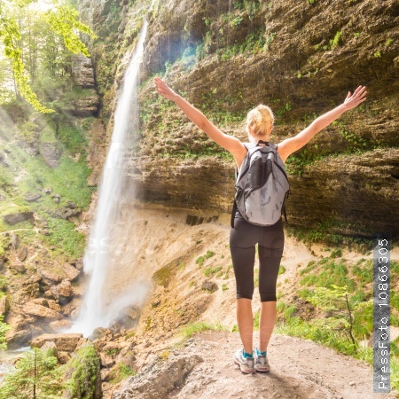Female hiker raising arms inhaling fresh air, feeling relaxed and free in beautiful natural environment under Pericnik waterfall in Vrata Valley in Triglav National Park in Julian Alps, Slovenia, stock photo