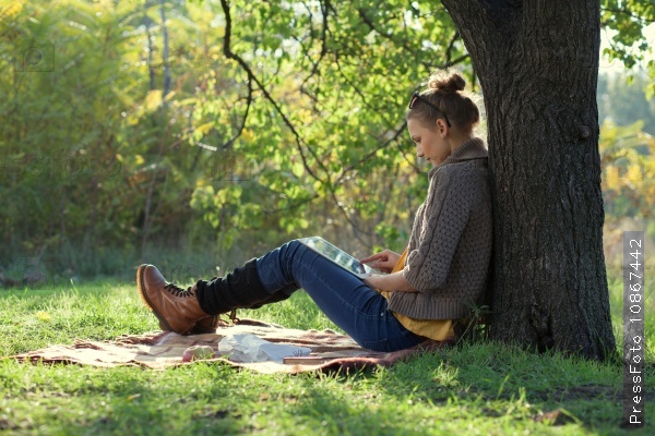Hipster girl leaning on a tree and using tablet computer