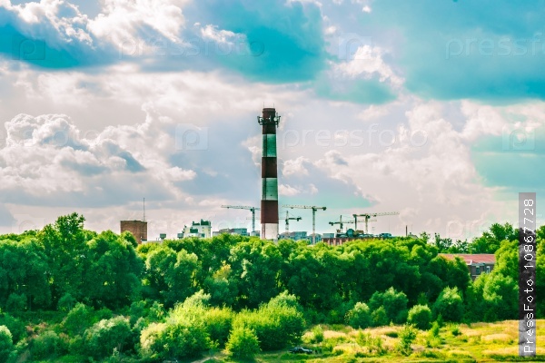 Chimney of a factory and construction site with cranes behind green trees. Industrial landscape with clouds in the sky and greenery.