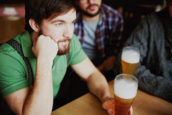 Young man with glass of beer watching football match in pub, stock photo