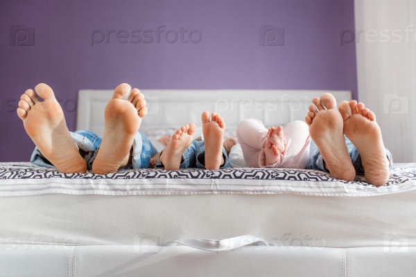 Funny family feet. Mother, father and two children lie on bed with white sheets; focus on feet.