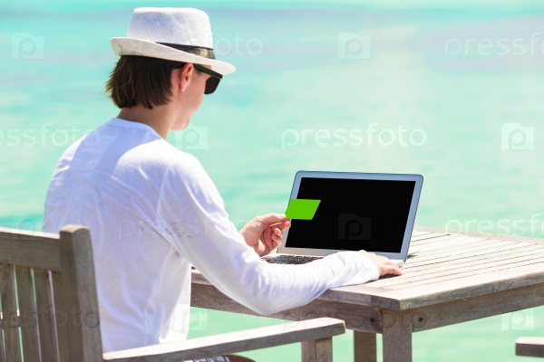 Young man working on laptop with credit card during tropical vacation, stock photo